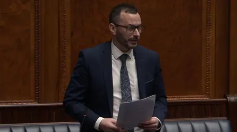 NI Assembly Timothy Gaston speaking in the assembly. He has short dark hair, wearing black glasses, a navy suit with white shirt and blue tie. He is holding a piece of paper with both hands.