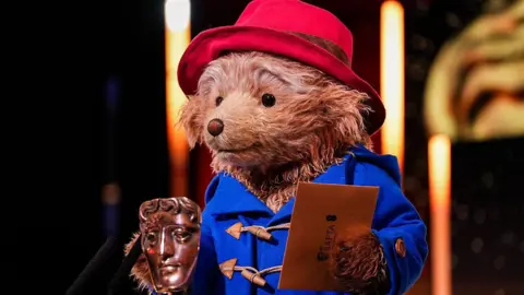 BAFTA via Getty Images A person in a bear costume holds a Bafta awards envelope in one paw, while there is an award in front of them. They wear a red hat and blue coat 