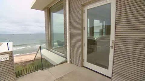 A modern building with a glass door labelled 'CLOSED' and large windows overlooking the beach and sea. The exterior features horizontal wooden slats, and there is a small terrace with glass railings. The sky is cloudy.