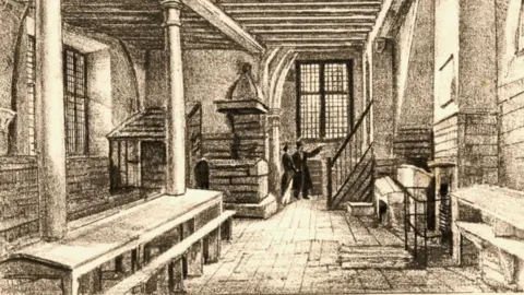 The Norris Museum A line drawing showing the interior of the Cromwell Museum in the mid-19th Century when it was a grammar school. It shows stone walls, two pillars holding up a wooden beamed roof, a fireplace, a long table with benches on either side and a man and woman at the far end under a window beside a small staircase.