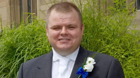 Merseyside Police Neil Doyle standing in front of a green bush outside of a church, wearing a wedding suit with white flower in the buttonhole and smiling.