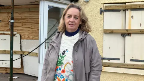 A woman wearing a grey coat and a white knitted jumper with a floral pattern on it stands outside a yellow house which has boards over its doors and windows.