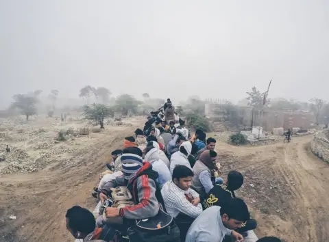 Shanu Babar The top of a train is crowded with men as it speeds along dirt roads.