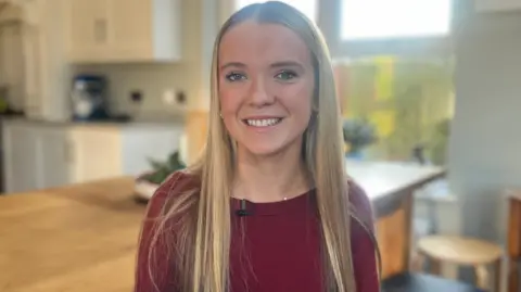 Emily smiling. She has very long straight blonde hair and is wearing a burgundy top.