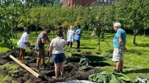 Victoria Holden A group of six adults around a large patch of mud being dug among rows of trees