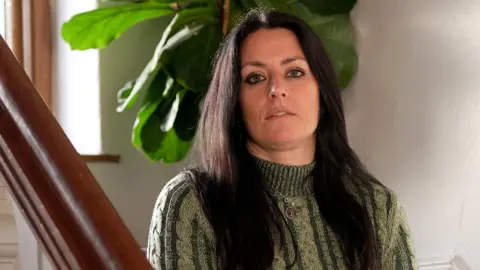 Rebecca has long dark hair and is wearing a green and cream polo neck sweater. She is sitting on the stairs with her hands on her lap and looking over the bannister into the camera. There is a plant with big green leaves behind her. She is not smiling.