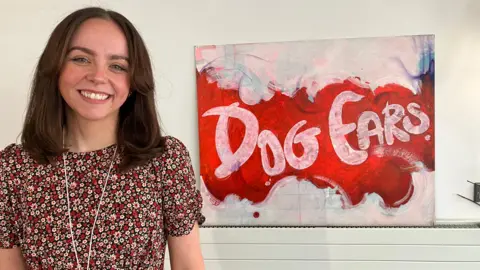 A woman with shoulder length brown hair, wearing a floral pattern top. She is smiling at the camera. There is a paining on the wall beside her with the words "Dog Ears" written on it.