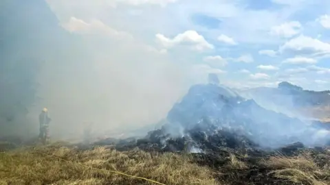 Norfolk Fire and Rescue Service Smoke billows from a smouldering straw fire in a field as a firefighter holds a hose with farm buildings in the background.