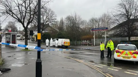 BBC Police tape is wound around a lamp-post on the left hand side of the image and goes across the road on the photo. A police car is on the right  of the photo with two people in police uniform next to it.