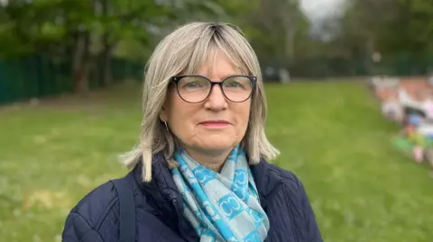 Janet Atkinson looks at the camera. She has shoulder length blonde hair and glasses and is wearing a two-tone blue scarf.