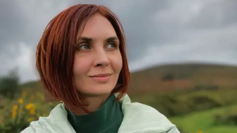A lady stood outdoors with hills and greenery in the background. She has short, reddish-brown hair and is wearing a light green jacket over a dark green top. They are holding the collar of the jacket with both hands. 
