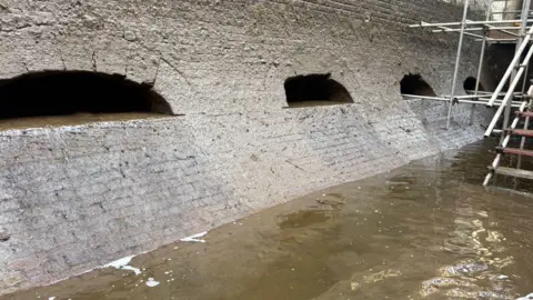 Canal and River Trust The inside wall of a canal lock is partially submerged, showing arched openings and scaffolding beside the water. Stairs can be seen in the back.
