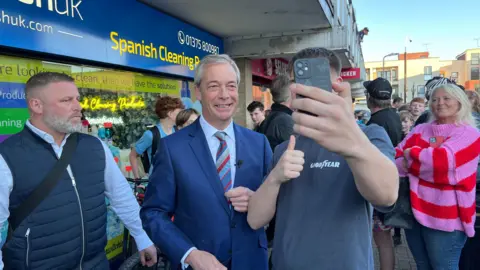 SIMON DEDMAN/BBC Nigel Farage posing for a selfie with a young man holding up an iphone which is covering up his face. Behind him are people who have come out on the street to see the Reform UK leader.  They are standing next to a shop with a sign saying spanish cleaning.