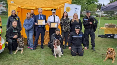 A group of 11 people, including police officers in uniform, stand and kneel on a stretch of grass, smiling at the camera. Three dogs are in the photo, with leads held by three police officers in uniform who kneel on the ground. An orange tent is behind them.