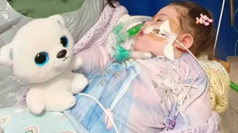 Two-year-old Alta Fixsler lies on a bed attached to medical equipment receiving treatment with a white teddy bear placed by her.