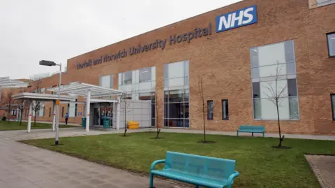 PA Media A bench is shown in the foreground next to grass and trees at the entrance of a building with a sign stating "Norfolk and Norwich University Hospital NHS" emblazoned in large lettering across its top section.