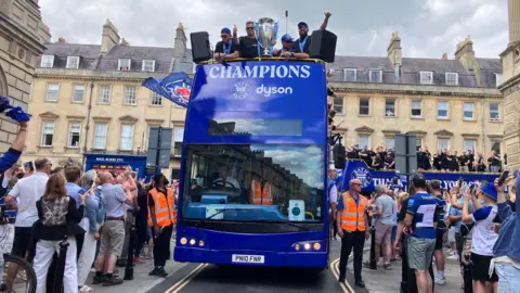A blue double decker bus with Champions written on the front alongside Bath Rugby's logo. There are several Bath Rugby players on the top deck with a big trophy. There are crowds on either side of the road by the bus, many of whom are in Bath Rugby clothing.