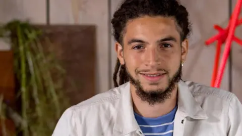Tayshan Hayden-Smith is seen beard wearing a white jacket, smiling in front of a wooden wall and red geometric sculpture.