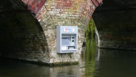 Facade of grey atm machine with screen, buttons on brick buttress with rippling water below 
