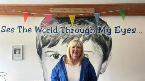 Head and shoulder view woman with medium-length blonde hair and a blue jacket standing in front of a giant poster showing a boy's head and some bunting above its banner line of "See The world Through my Eyes" 