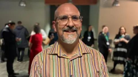 Image if Simon. He has a bald head and a grey goatee beard. He is wearing glasses and a multi-coloured stripped shirt. There are people in the background.