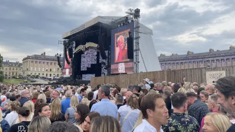 A large crowd of people standing near the front of the stage at the Royal Crescent in Bath. It is a cloudy day and everyone is stood around chatting as the music has not started yet. 