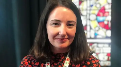 Emer O'Donnell is in a red and black top and is wearing a lanyard. 
She has should-length black hair. 