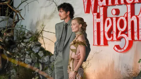 Getty Images Margot Robbie and Jacob Elordi pose before a moody “Wuthering Heights” backdrop of red lace‑patterned letters, rocks, and branches; one wears a grey suit, the other an ornate gold‑and‑green outfit.