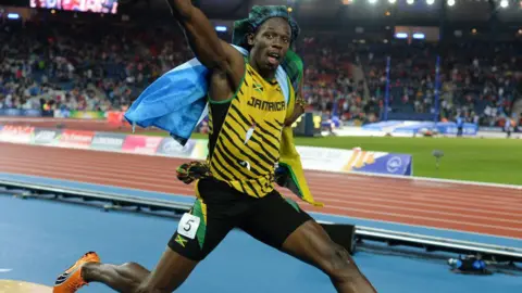 SNS Usain Bolt świętuje sukces na Igrzyskach Wspólnoty Narodów w 2014 r. z zapiętym króliczkiem i flagą.