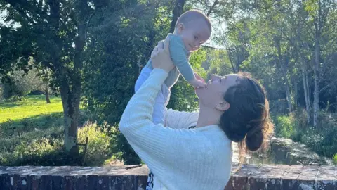 NHSBT A woman stands outdoors holding a baby near a brick wall, with a river, trees, and sunlight visible in the background.