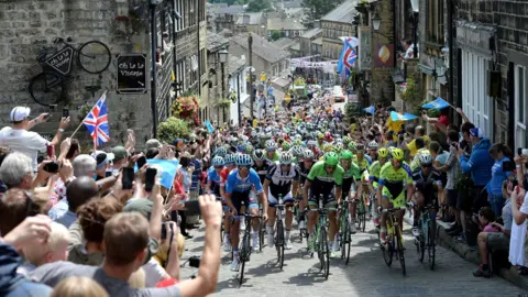 PA Media Hundreds of cyclists ride up a cobbled street lines with supporters. There are British flags lining the historic street.