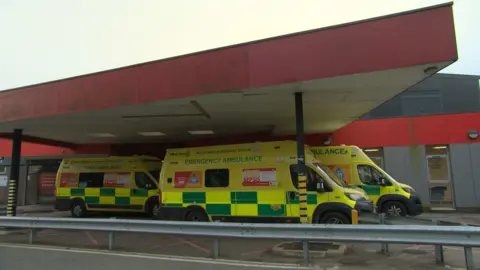 The A&E department at Macclesfield hospital. Three ambulances are parked under a canopy. The building behind is grey and red. 