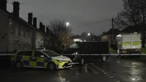 A suburban street on a rainy evening. It is cordoned off by police tape. A police car, a police van are visible, as is a black van. 
