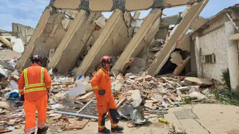 Two firefighters dressed in orange standing in front of a damaged building and pile of rubble.