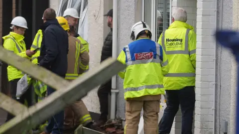 PA People in yellow high vis jackets are outside a house. Some are wearing hard hats. One the back of one jacket states safety officer and on the back of another jacket says Housing Executive.