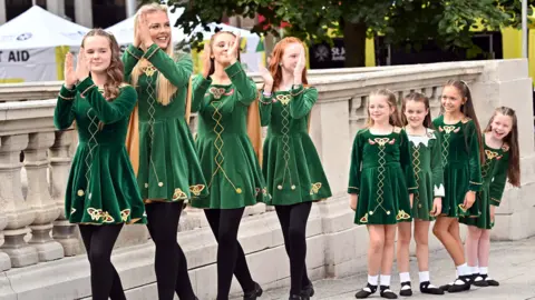 Irish dancers from the Reilly School of Dancing Belfast.
There are 8 dancers all wearing green dancing dresses.
The first 4 are older and have their hands raised to clap. the second 4 are young girls watching on