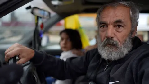 BBC Abu Ali sits in the front seat of his car, hand on the steering wheel. A child and presumed relative sits in the passenger seat next to him.