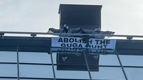 Two men dressed as seabirds are on the roof of a building with their arms outstretched. They are holding a sign which reads "abolish the guga hunt". The building has large glass panels immediately below them. They are in front of what looks like a ventilation tower. 