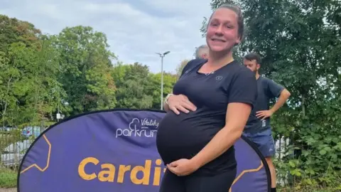 Zoe Jacobs Zoe holding her baby bump in front of a Parkrun sign