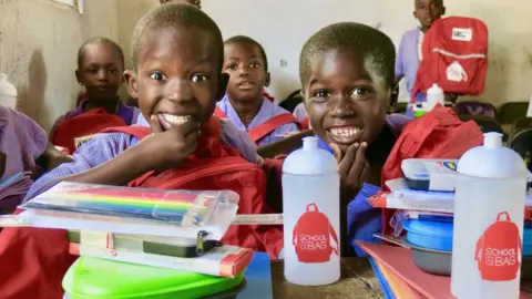 School in a Bag Two boys smiling in a classroom holding new red rucksacks with school supplies