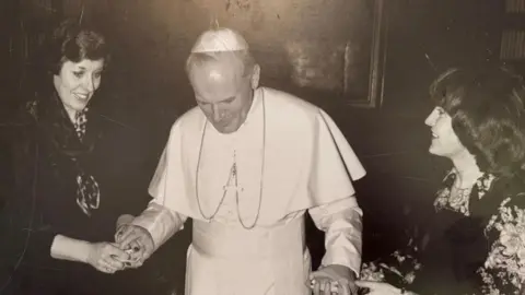 Two women with 1980s style hair hold either hand of Pope John Paul II, dressed in his white papal gown and skullcap. 