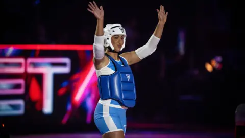 Keeva wearing blue and white safety gear, she is holding her hands in the air