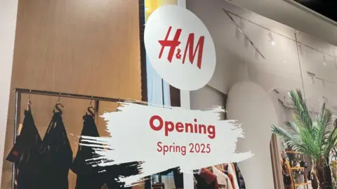 A H&M sign with "Opening Spring 2025" written underneath. The words are in red on a white background superimposed on a picture of a clothes store.