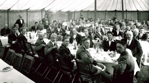 Essex County Cricket Club Smartly dressed men face the camera for a picture taken in black and white. They are sitting at long tables inside a large white tent. It appears they are either about to enjoy a pre or post-match meal.