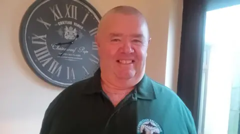 a man wearing a Services Club Ballymena branded poloshirt, smiles at the camera