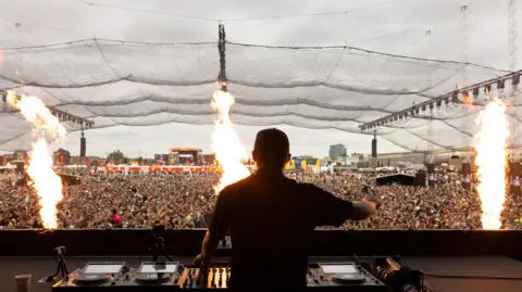 Dom Dolla has his back to the camera and is case in shadow. He's wearing a T-shirt and cap. In front of him is a long desk with DJ decks and cameras on little tripods. Beyond the desk is a sea of people stretching out into the distance. Three jets of flame blast from the front of the stage as part of a pyrotechnic display. 
