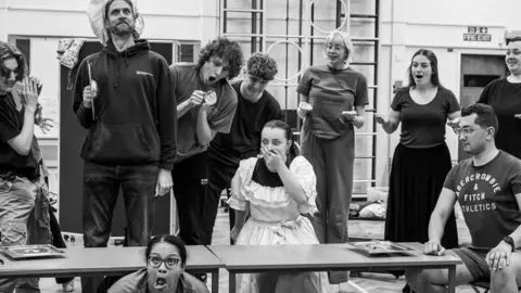 Cat Humphries 10 actors rehearsing a scene from The Little Mermaid. One is on the floor pulling a face, a woman looks shocked, with her hand on her mouth, one man is looking out with a hat, the rest are looking down at the women in shock. The image is black and white and they are in a school hall with equipment behind on the wall. 