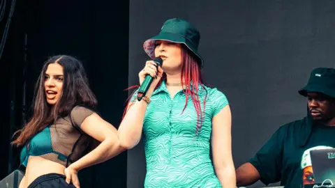 Three women and a man are performing on a festival stage. The woman in the centre has on a turquoise jumpsuit with wave patterns on and a bucket hat. She has long red hair and is singing into a microphone. The two women to her side are wearing bikinis to match her dress and black trousers. A man is mixing at decks in the background. 