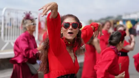 PA Media A woman dressed in a red dress with heart-shaped sunglasses dances. More people in red are dancing behind her.