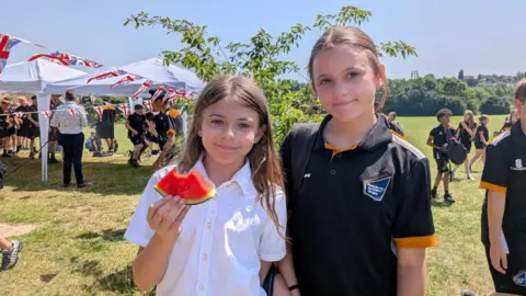 Beacon Hill Academy Two young schools girls pictured out on the school playing field. The girl on the left holds up a partially eaten watermelon slice while the girl on the right smiles with her arm around her friend.